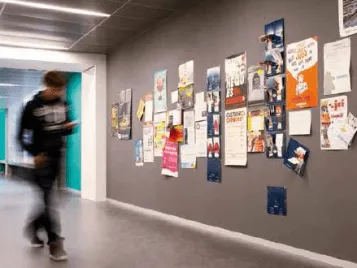 A corridor with grey flooring and a grey wall adorned with pieces of paper, a person is walking through